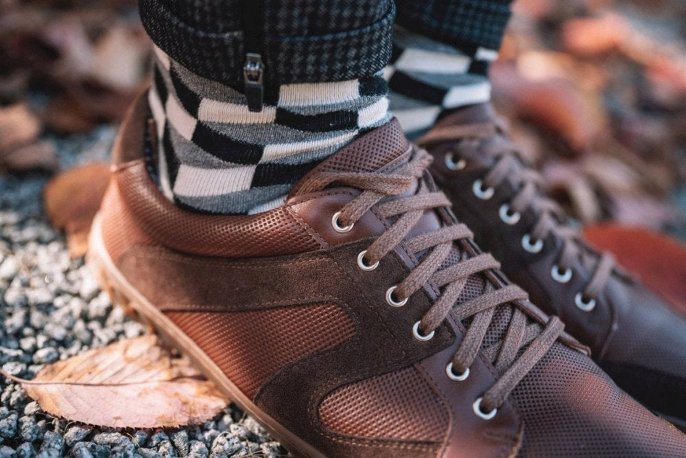 Eine Nahaufnahme eines Paars RIQO Brown-Sneaker von ZAQQ mit Schnürsenkeln, getragen mit schwarz-weiß gestreiften Socken. Die Schuhe ruhen auf einer mit Herbstblättern bestreuten Kiesoberfläche und bieten außergewöhnlichen Komfort und ein atmungsaktives Erlebnis bei jedem Schritt.