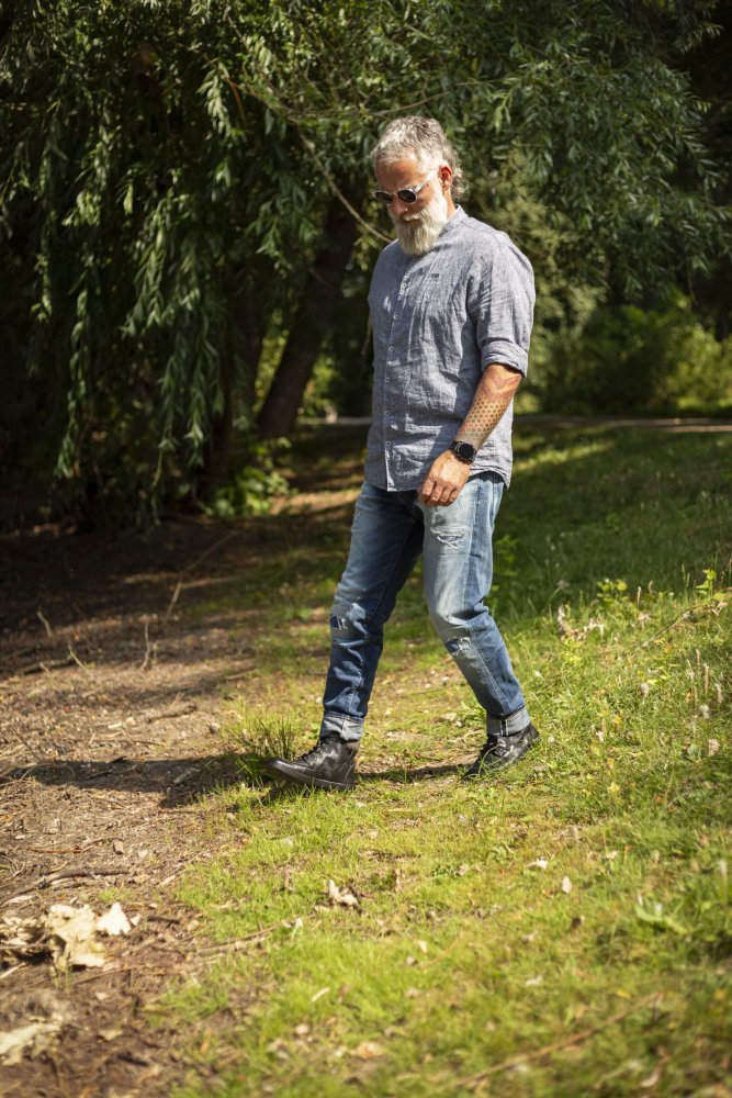 Ein bärtiger Mann mit Sonnenbrille, grauem Hemd und Jeans schlendert durch einen schattigen Parkbereich und stellt inmitten des üppigen Grüns und der Bäume seine Lieblingssneakers „ZAQQ ROQA Black“ zur Schau.