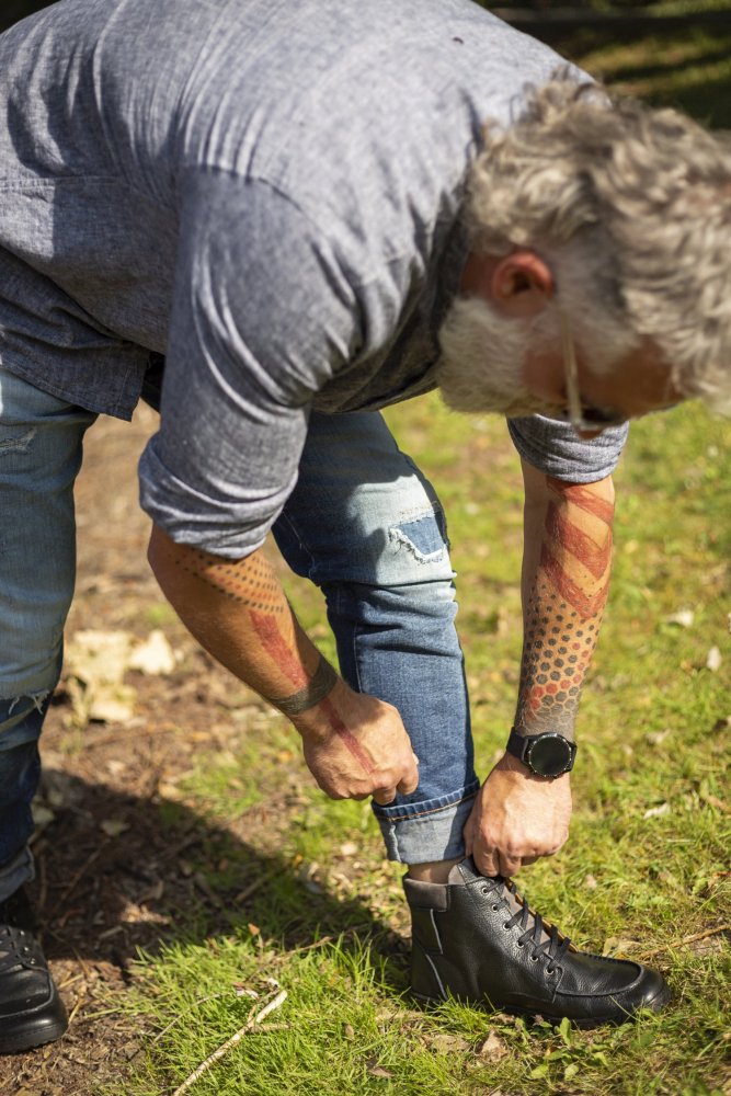 Ein Mann mit grauem Haar und Bart rückt den Saum seiner Jeans zurecht und enthüllt einen tätowierten Arm. Er trägt ein graues Hemd und schwarze ROQA-Stiefel von ZAQQ, komplett mit einem dezenten Reißverschluss. Die Szene spielt im Freien auf einer Wiese, durch die gesprenkeltes Sonnenlicht fällt.