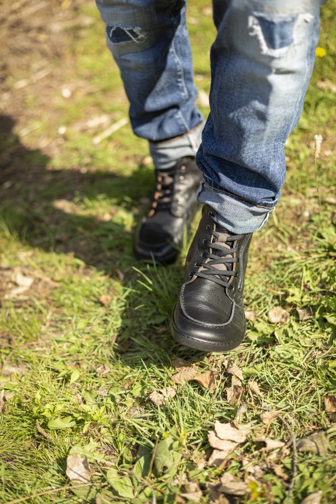 Eine Person, die ZAQQs schwarze ROQA-Lederstiefel mit dezentem Reißverschluss und zerrissene Bluejeans trägt, geht auf einem Grasweg mit verstreuten Blättern und Zweigen, die einen Schatten auf den Boden werfen.