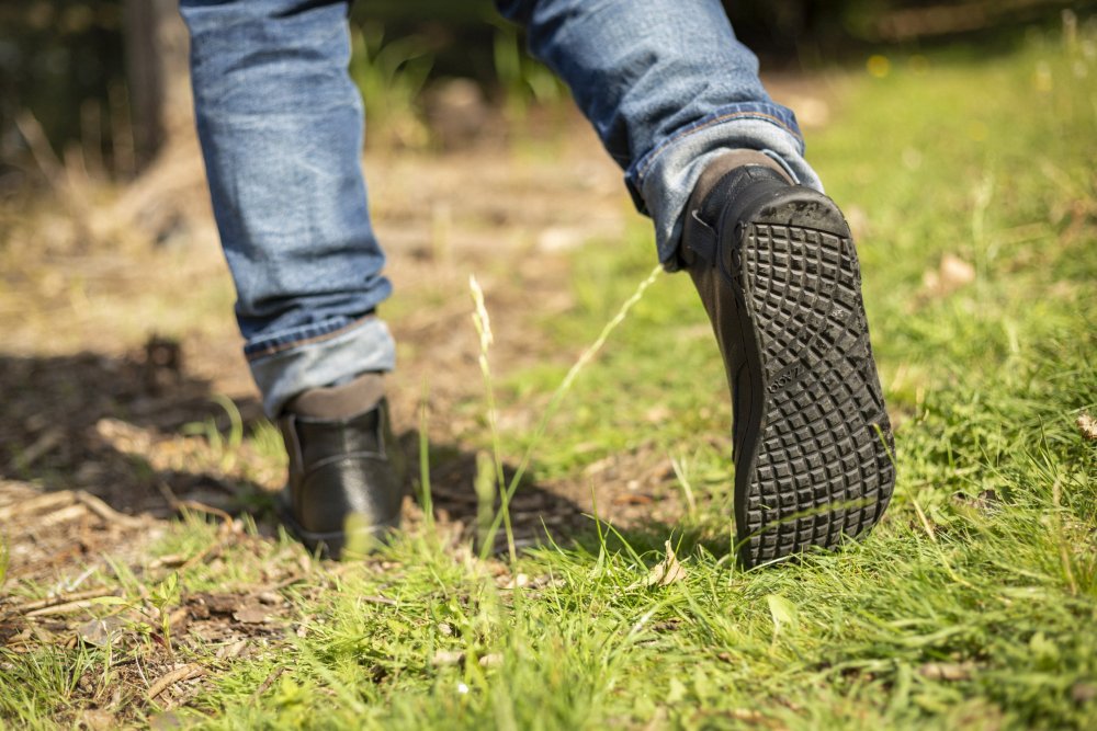 Nahaufnahme einer Person, die auf einem Grasweg läuft. Sie trägt Jeans und die schicken ROQA Black Sneakers von ZAQQ. Der Fokus liegt auf der Sohle eines Schuhs, die ihr strukturiertes Muster offenbart. Der Hintergrund ist eine verschwommene, natürliche Umgebung.