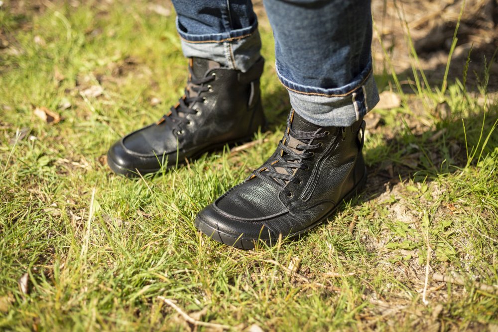 Eine Person in hochgekrempelten Bluejeans und robusten schwarzen Lederstiefeln ROQA Black von ZAQQ steht auf einer Rasenfläche, auf der Zweige und trockene Blätter verstreut liegen. Die geschnürten ROQA Black-Stiefel werden durch robuste Reißverschlüsse ergänzt, was darauf hindeutet, dass sie für Outdoor-Aktivitäten geeignet sein könnten.