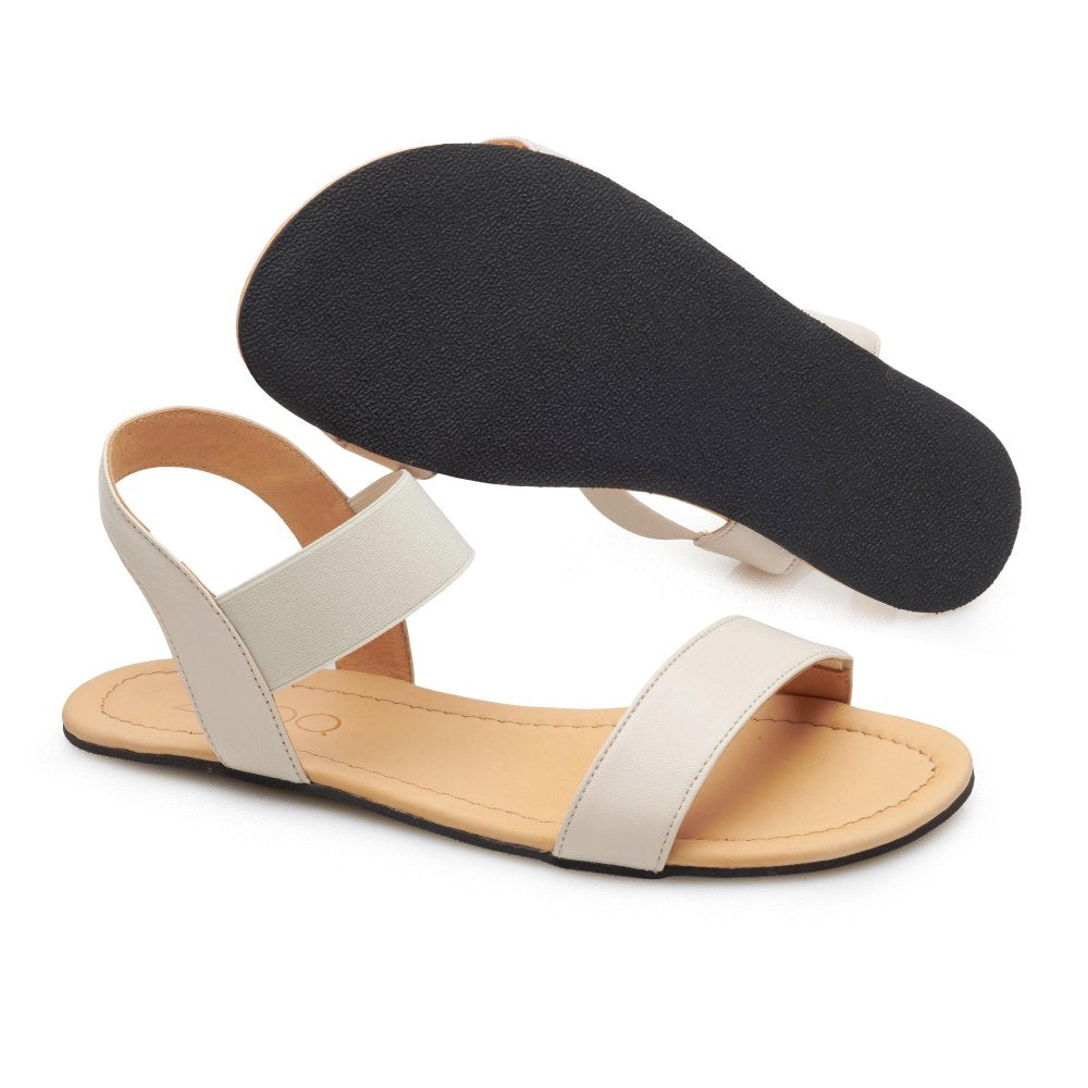 Das ZAQQ SLIQ Beige ist ein Paar flache Barfuß-Sandalen mit beigen Knöchelriemen und schwarzer Sohle, die für ihren außergewöhnlichen Komfort bekannt sind. Eine Sandale steht aufrecht und betont das Design, während der andere Winkel die Sohle einzigartig hervorhebt.
