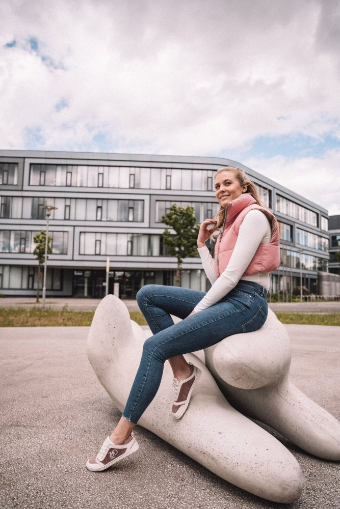 Eine Person in einer rosa Weste und Jeans sitzt auf einer großen abstrakten weißen Skulptur in einer städtischen Umgebung und hebt ihre stylischen ZAQQ SQILL Rose-Sneaker aus veganer Mikrofaser hervor, mit einem modernen Gebäude im Hintergrund und einem bewölkten Himmel darüber.