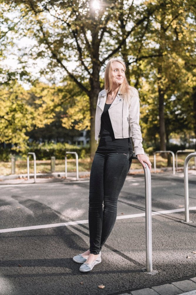 Eine Frau mit langen blonden Haaren, bekleidet mit einer leichten Jacke und schwarzen Hosen, lehnt an einem Metallgeländer. Sie steht auf einem gepflasterten Weg und trägt ein elegantes Paar SUQAR Grey-Schuhe von ZAQQ aus weichem Nappaleder, während das Sonnenlicht durch die Bäume hinter ihr fällt.