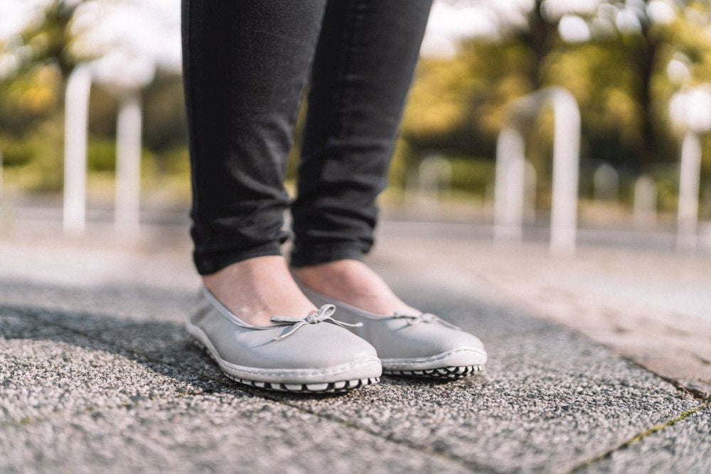 Eine Person in SUQAR Grey Ballerinas von ZAQQ und schwarzen Hosen steht auf einem strukturierten Steinweg. Der Hintergrund ist verschwommen mit Andeutungen von Grün und weißen Pfosten.