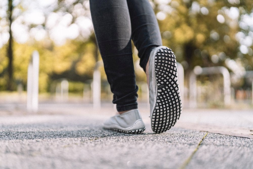 Nahaufnahme einer Person, die auf einem gepflasterten Weg geht und schwarze Jeans und die trendigen SUQAR Grey-Sneaker von ZAQQ trägt, deren gemusterte Sohle Barfußschuhen nachempfunden ist. Der Hintergrund ist verschwommen und hebt Bäume und sanftes Sonnenlicht hervor, was eine ruhige Parklandschaft suggeriert.