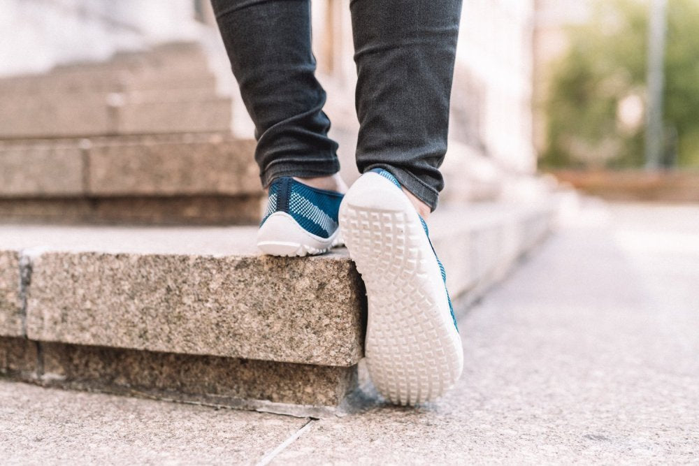 Eine Person in Jeans und blauen Sneakers, das SUQAR Mare von ZAQQ, läuft im Freien Steintreppen hinunter. Der Fokus liegt auf den Schuhen und Stufen, vor einem verschwommenen grünen Hintergrund.