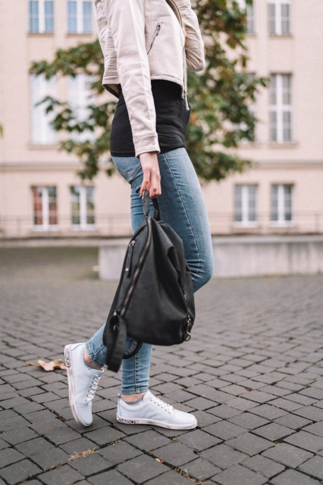 Eine Person trägt blaue Jeans, die ZAQQ TAQQ Nappa Lightblue Sneakers mit flexibler Sohle für mehr Komfort und eine leichte Jacke und spaziert mit einem schwarzen Rucksack in der Hand über eine gepflasterte Fläche. Im Hintergrund sind ein Gebäude mit großen Fenstern und ein Baum zu sehen.