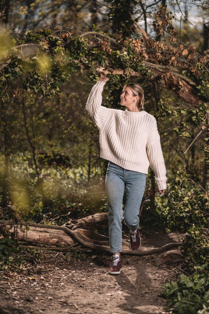 Eine Person in einem weißen Pullover und Jeans geht auf einem Waldweg und trägt bequeme TREQ Red Waterproof-Schuhe von ZAQQ. Sie hebt einen Ast über ihren Kopf und genießt den sonnigen Tag im Freien.