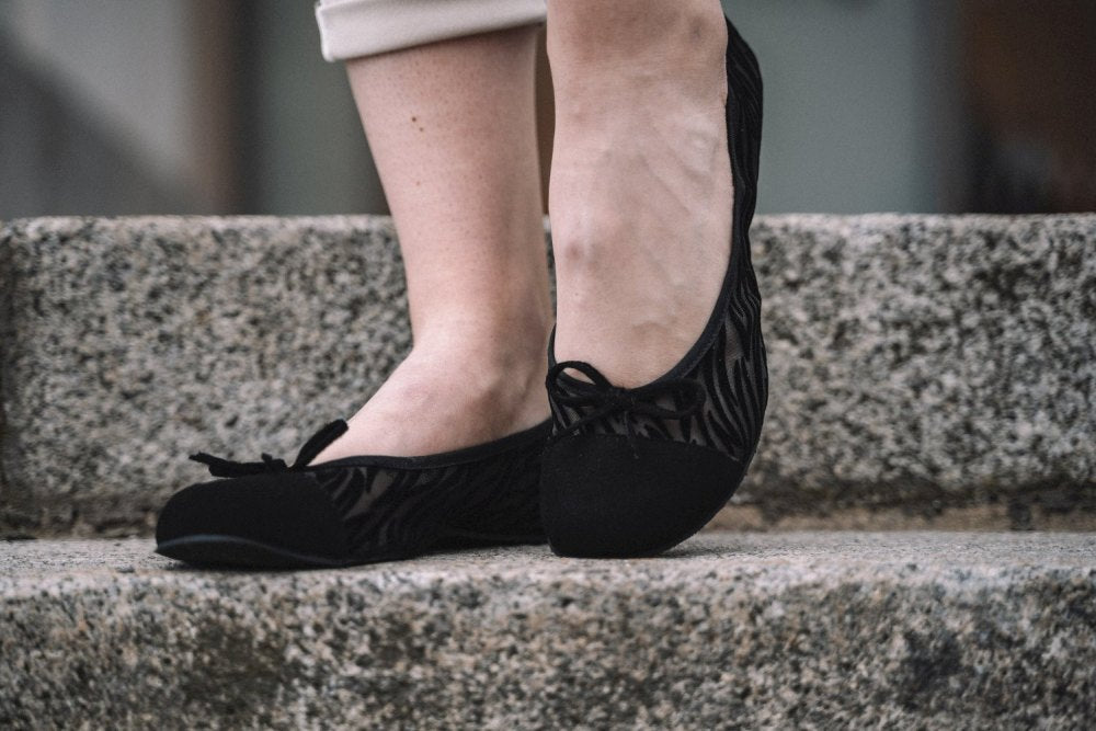 Nahaufnahme einer Person, die die TWIST Sling Black Flats von ZAQQ mit dezentem Muster und flexibler Sohle trägt und auf Steinstufen steht. Die Schuhe, die mit kleinen Schleifen an den Zehen verziert sind, passen perfekt zu hellen Hosen für einen schicken und bequemen Look.