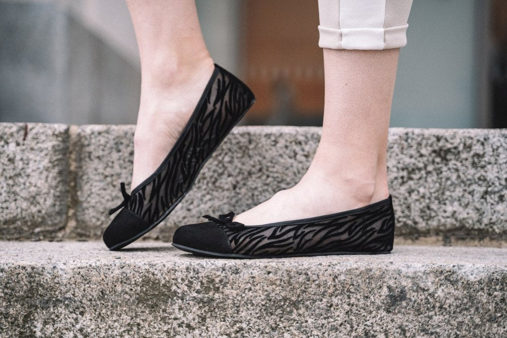 Eine Person in den ZAQQ TWIST Sling Black Ballerinas steht auf einer Steintreppe. Mit einer flexiblen Sohle und einer weißen Hose kombiniert, werden die Füße stilvoll in Szene gesetzt.