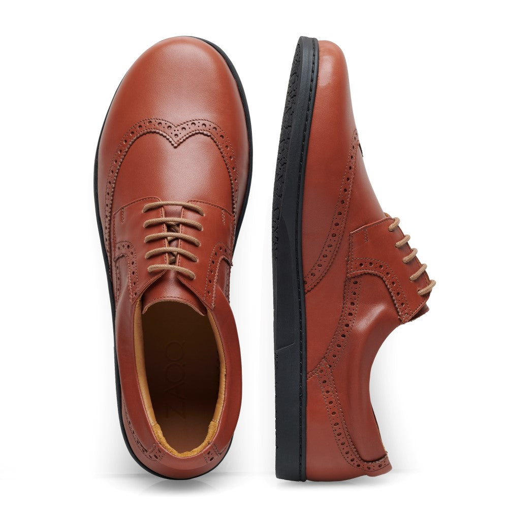 Die BRIQ Brogue Cognac Schuhe von ZAQQ vereinen klassischen Stil mit Komfort und sind somit der ideale bequeme Businessschuh. Der linke Schuh ist geschnürt und von oben zu sehen, während der rechte Schuh von der Seite zu sehen ist. Beide Schuhe haben schwarze Sohlen und ein poliertes Finish.