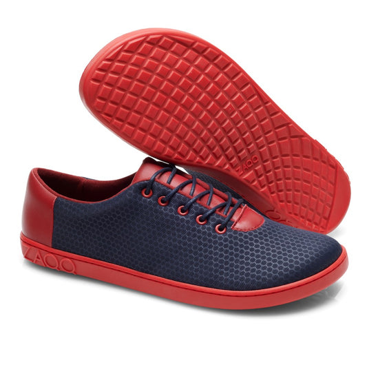 Ein Paar QARO Navy Red von ZAQQ, mit einem stylischen marineblauen Mesh-Obermaterial mit Wabenmuster und leuchtend roten Akzenten an der Kautschuksohle, den Schnürsenkeln und der Ferse. Die Schuhe werden so präsentiert, dass ein Schuh flach liegt, während der andere aufrecht steht, was die auffällige rote Sohle betont.