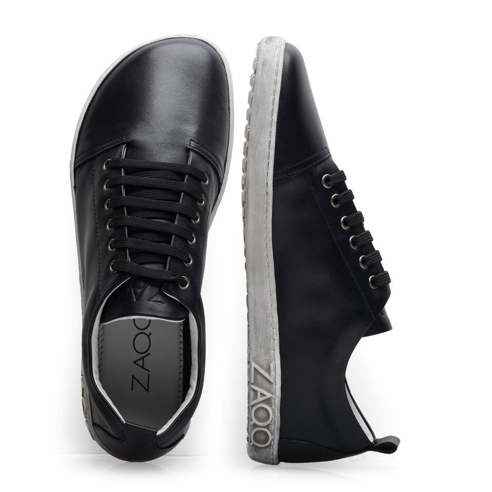 Ein Paar elegante TAQQ Nappa Black-Sneaker aus glänzendem schwarzem Leder mit weißen Sohlen werden aus zwei Blickwinkeln präsentiert: einmal von oben, wodurch die Schnürsenkel hervorgehoben werden, und einmal von der Seite, mit dem in die Ferse geprägten Markennamen ZAQQ.