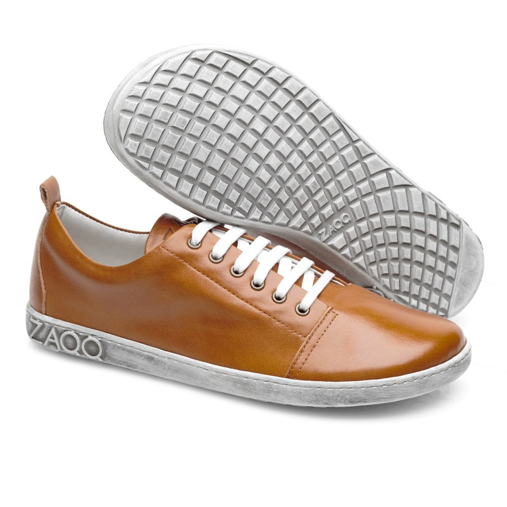 Ein Paar TAQQ Nappa Brown-Sneaker in Braun mit weißer Sohle und Schnürung. Ein Schuh steht aufrecht, während der andere angewinkelt ist, um die flexible Barfußsohle zu präsentieren. Der Markenname ZAQQ ist elegant auf der Seite jeder Sohle eingeprägt.